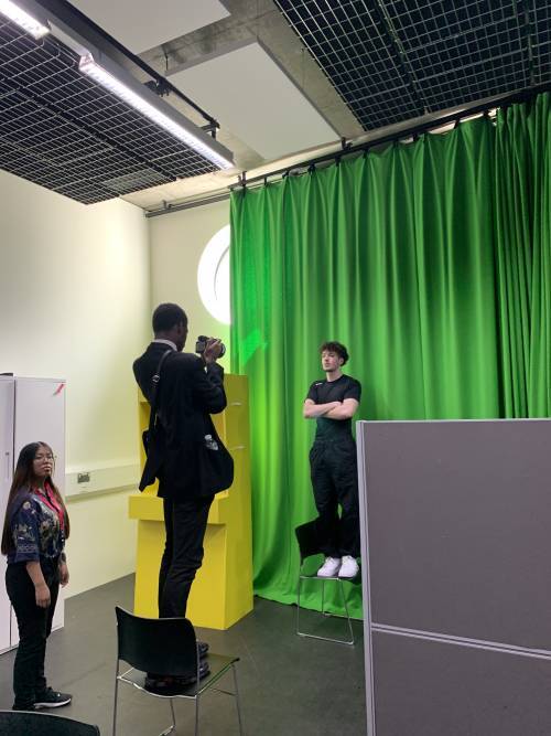 Two men stand opposite each other on chairs while one takes a photograph of the other against a green screen